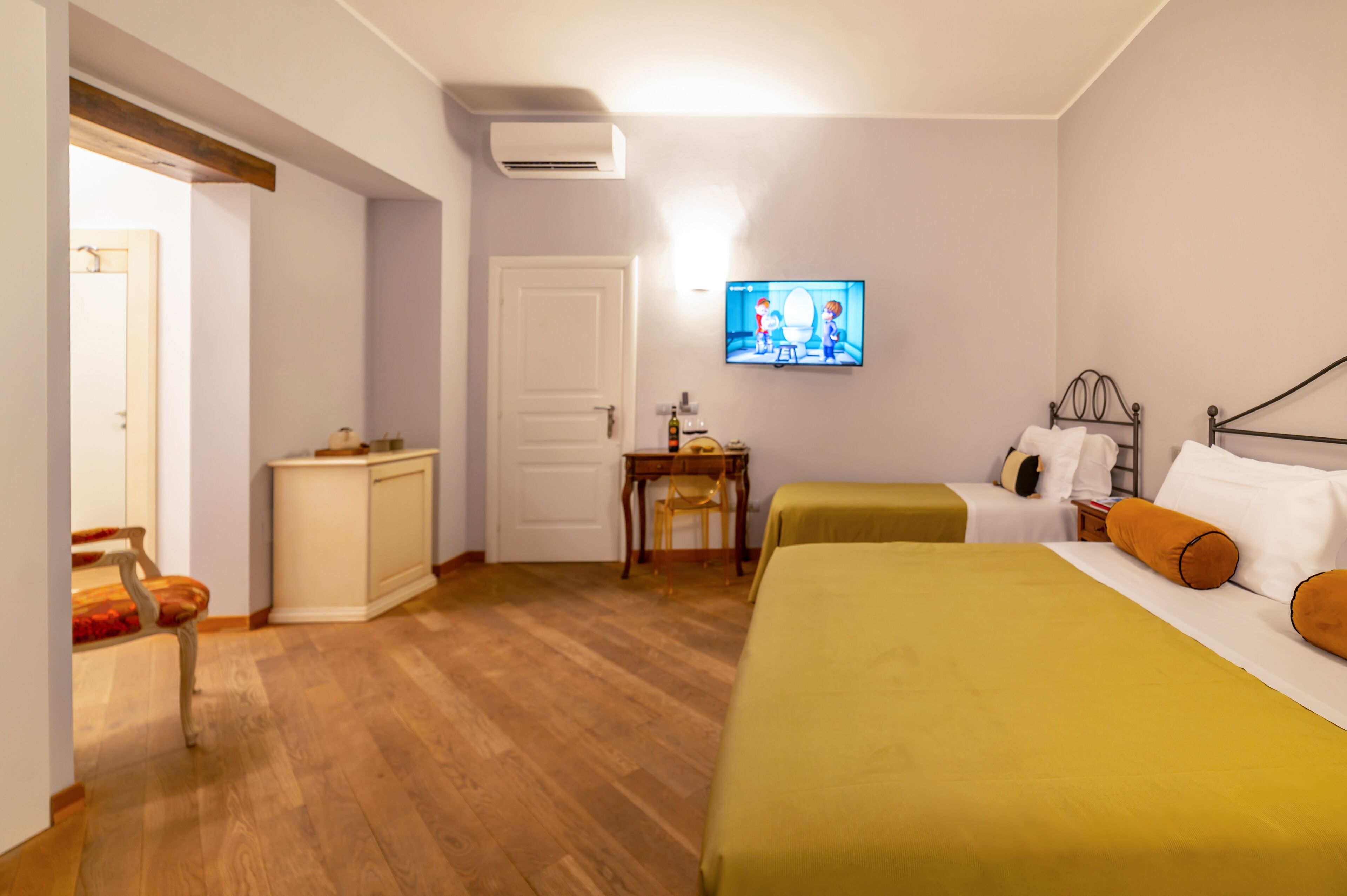 A hotel room with a wooden floor, a bed with a yellow bedspread, a television mounted on the wall, a desk with a chair, and a door.