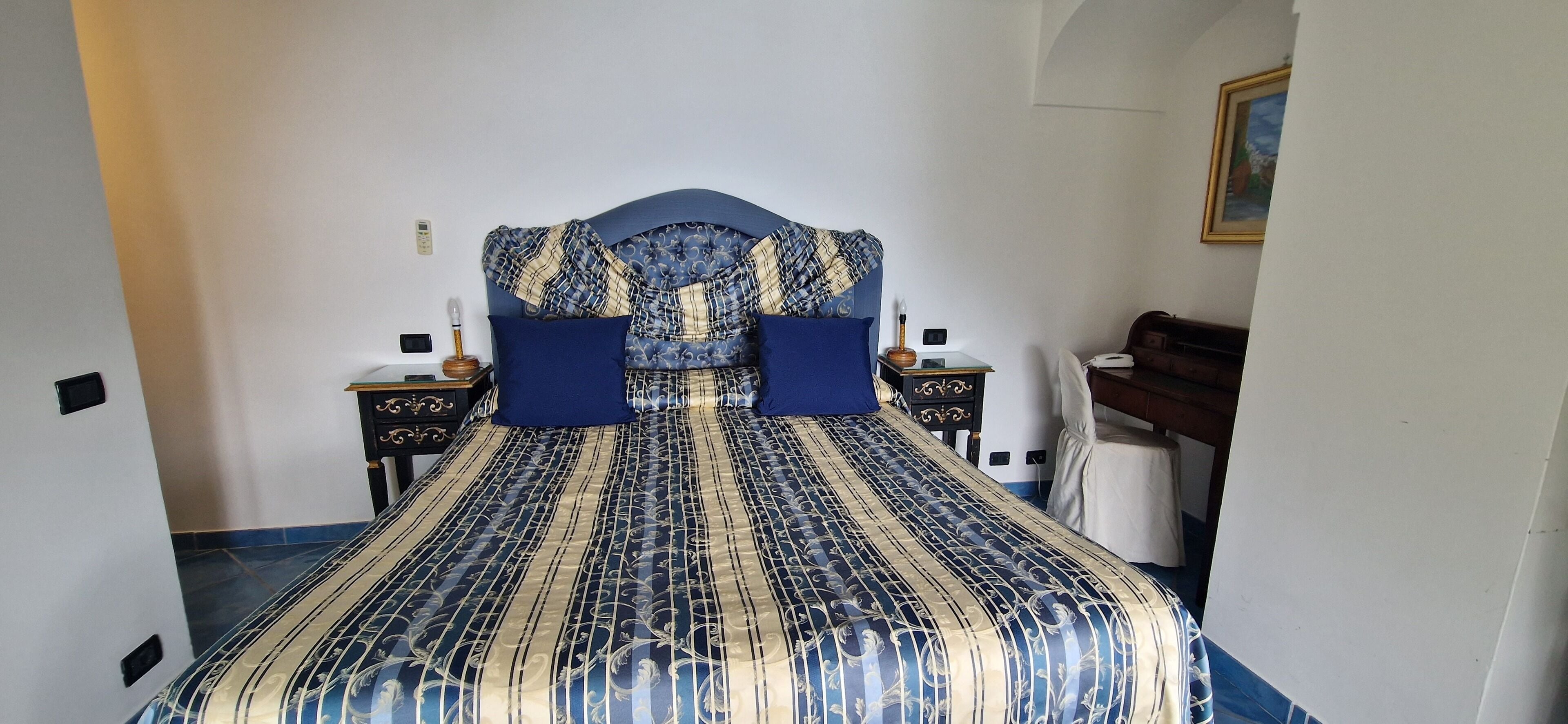 Een net opgemaakt bed met een blauw-en-goud geborduurde sprei, twee bijpassende kussens, en een hoofdbord met een blauw-wit geruit patroon.