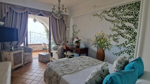 A bedroom with a bed, a desk, a chair, a TV, a balcony with curtains, and a wall with a leaf pattern. - La Conca Azzurra (Conca dei Marini)