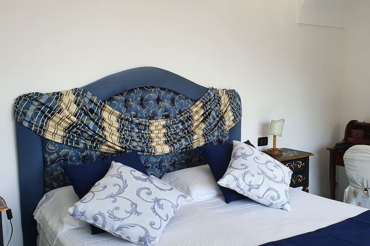A neatly made bed with a blue headboard, white and blue patterned pillows, and a blue bedspread. There is a nightstand with a lamp and a small table with a lamp. A painting is on the wall, and a chair is partially visible.