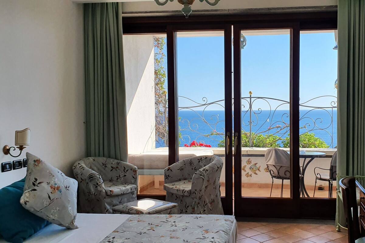 A hotel room with a bed, two armchairs, a small table, and a balcony with a view of the sea.