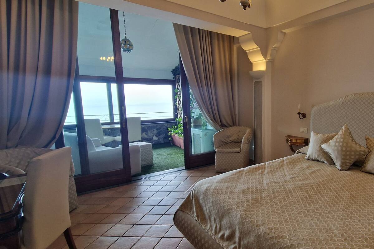 A bedroom with a large bed, a chair, a desk, and a view of the outdoors through a sliding door.