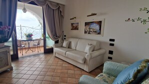 A living room with a beige sofa, a small table with a vase of flowers, a balcony with a table and chairs, and two framed pictures on the wall. - La Conca Azzurra (Conca dei Marini)