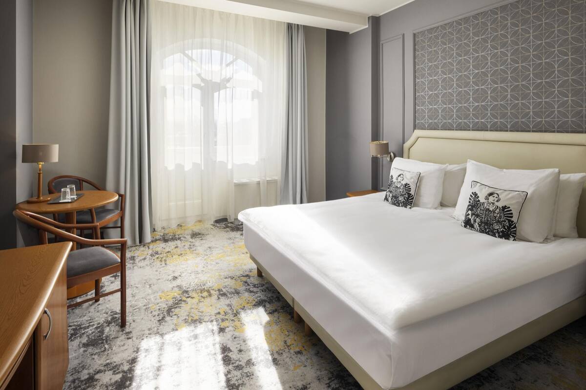 Ein Hotelzimmer mit einem großen Bett, einem Schreibtisch mit Stuhl, einer Lampe und einem Fenster mit durchsichtigen Vorhängen.