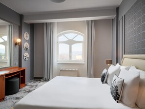 Habitación de hotel con cama, escritorio, silla, ventana y lámpara de pared.