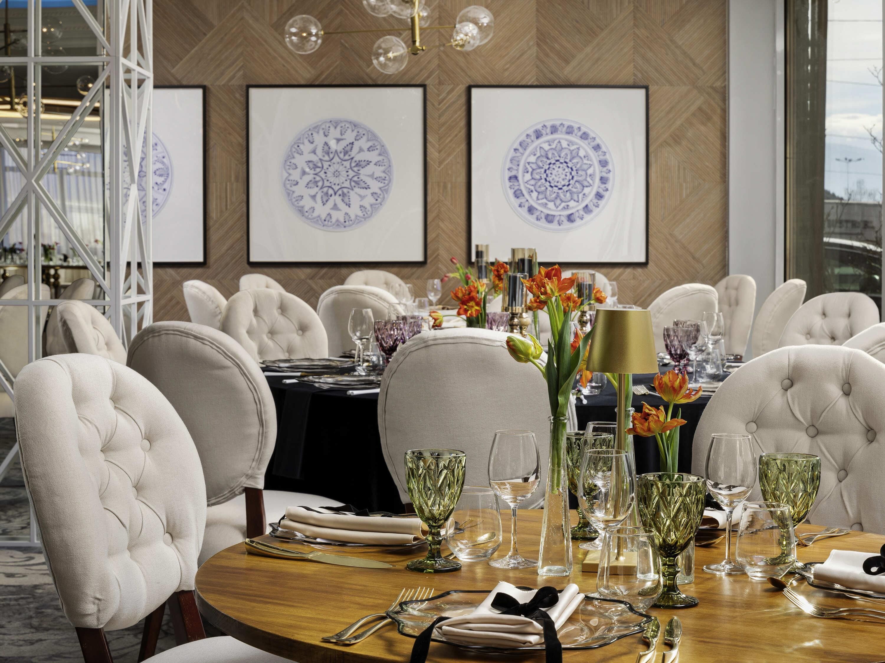 Un comedor con una mesa redonda dispuesta para seis personas, rodeada de sillas tapizadas. Se observan adornos enmarcados en la pared sobre la mesa, y un candelabro colgando del techo.