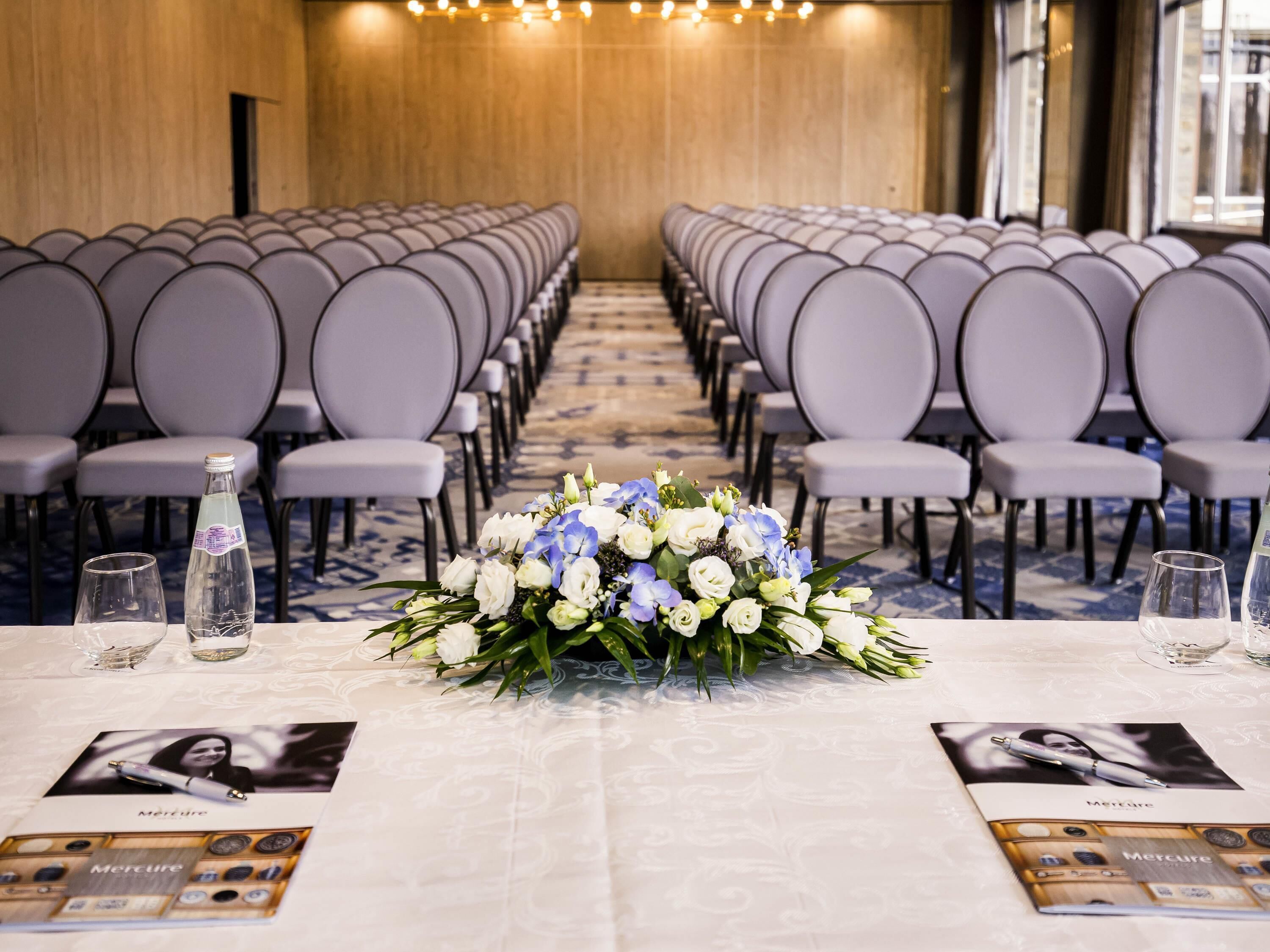Une salle de conférence avec des rangées de chaises, une table ornée d’un centre de table floral et des brochures disposées sur la table.