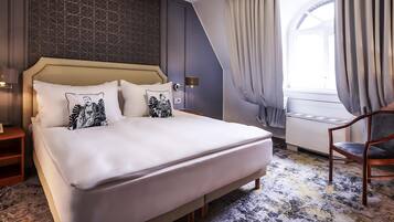 A neatly made bed with a headboard, a bedside table with a lamp, and a chair in a room with a patterned carpet and a window with curtains.