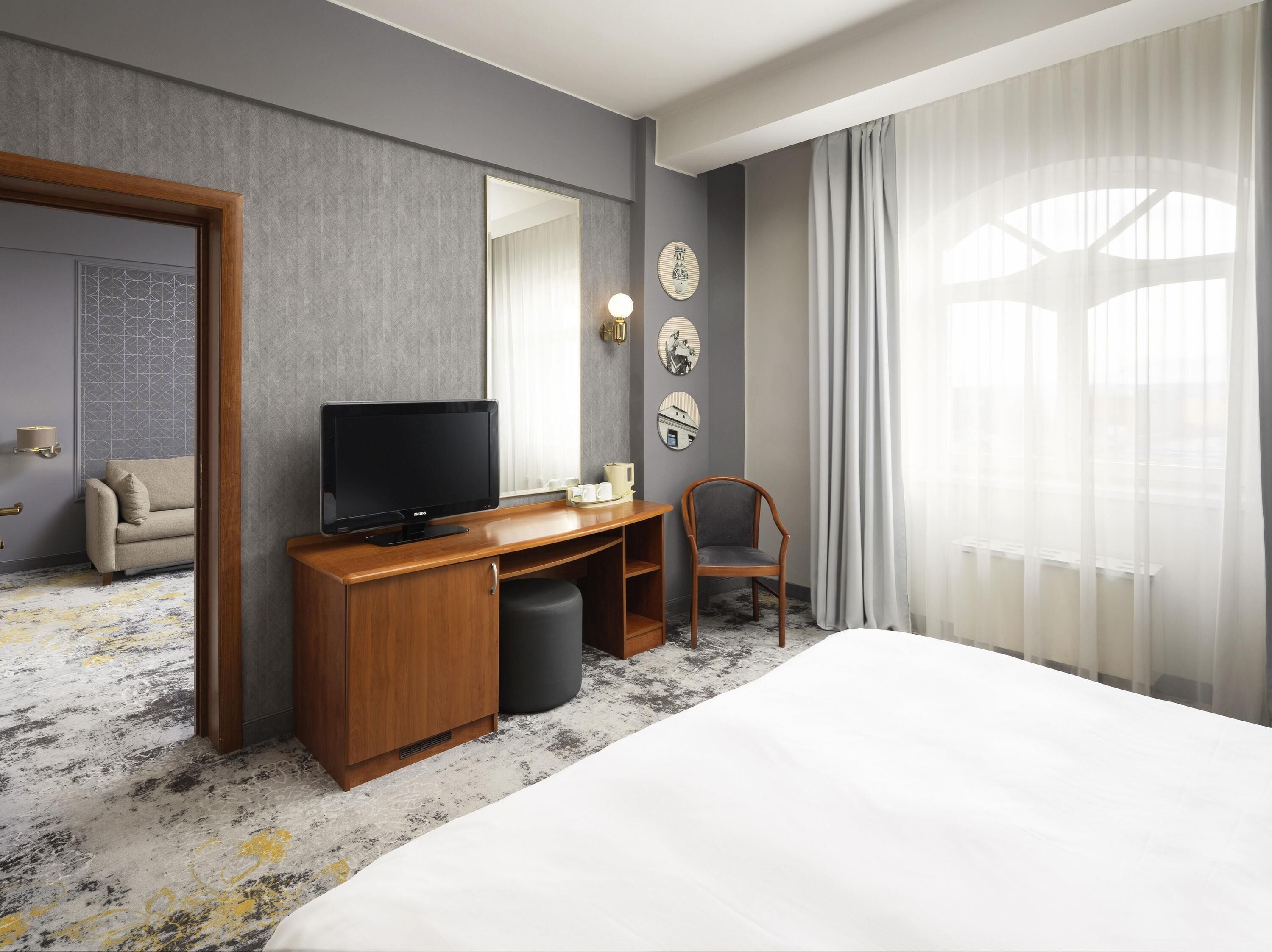 Une chambre d’hôtel avec un lit, un bureau en bois avec une télévision, une chaise et une grande fenêtre avec des rideaux.