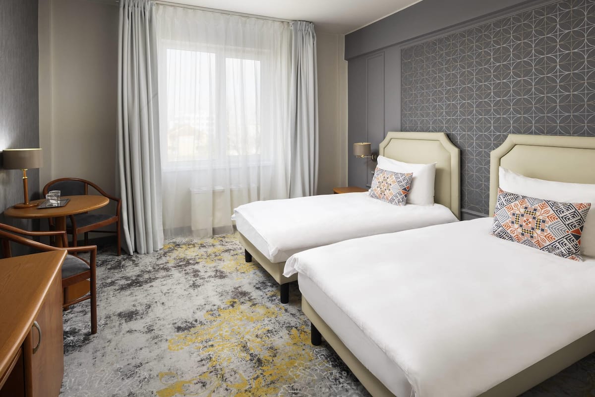 Ein Hotelzimmer mit zwei Betten, einem Schreibtisch mit Stuhl, einer Lampe und einem Fenster mit durchsichtigen Vorhängen.