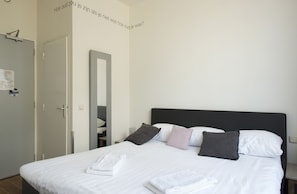 Una cama bien hecha con sábanas blancas y algunos almohadones, una puerta a la izquierda y un espejo en la pared.