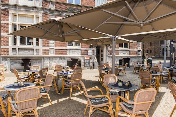 Un café al aire libre con sillas y mesas de mimbre, sombrillas grandes y un edificio de ladrillo al fondo.