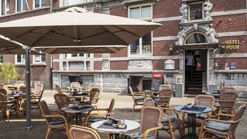 Un café al aire libre con mobiliario de mimbre y sombrillas, frente a un edificio de ladrillo histórico con entrada visible a un hotel.