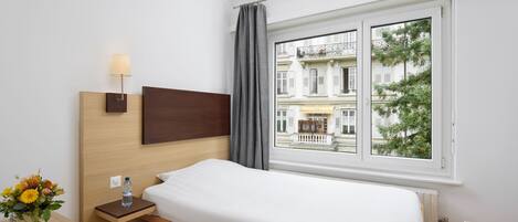 Ein Hotelzimmer mit Bett, Nachttisch, Lampe und Fenster mit Blick auf Gebäude und Bäume.