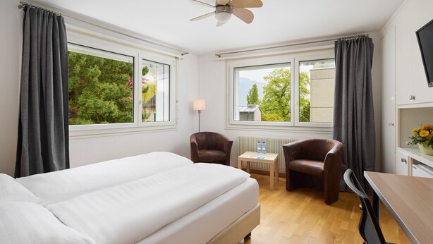 A hotel room with a bed, two chairs, a table, a lamp, and a ceiling fan. - Hôtel Bon Port (Montreux)