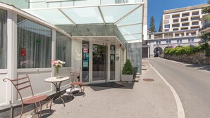A hotel entrance with a glass canopy, a small outdoor seating area with two chairs and a table, and a sign indicating the hotel name. - Hôtel Bon Port (Montreux)