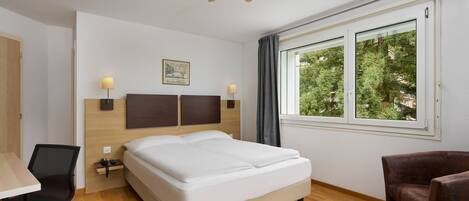 Habitación de hotel con cama, escritorio, silla, ventilador de techo, ventana con vistas a árboles y lámpara de pared.