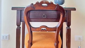 Una silla de madera con asiento y respaldo acolchados, una mesa con lámpara y un cuadro en la pared.