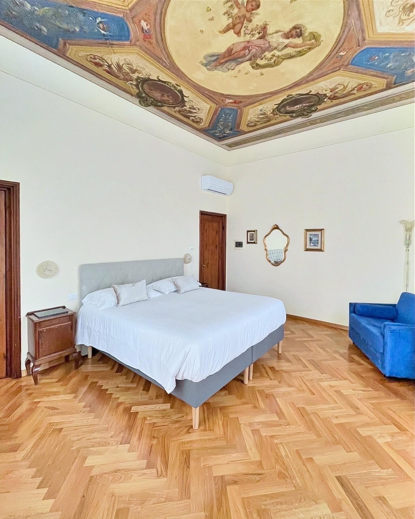 Ein Schlafzimmer mit einem großen Bett, Holzboden, Wandmalerei an der Decke, einem blauen Sofa und einem Nachttisch aus Holz.
