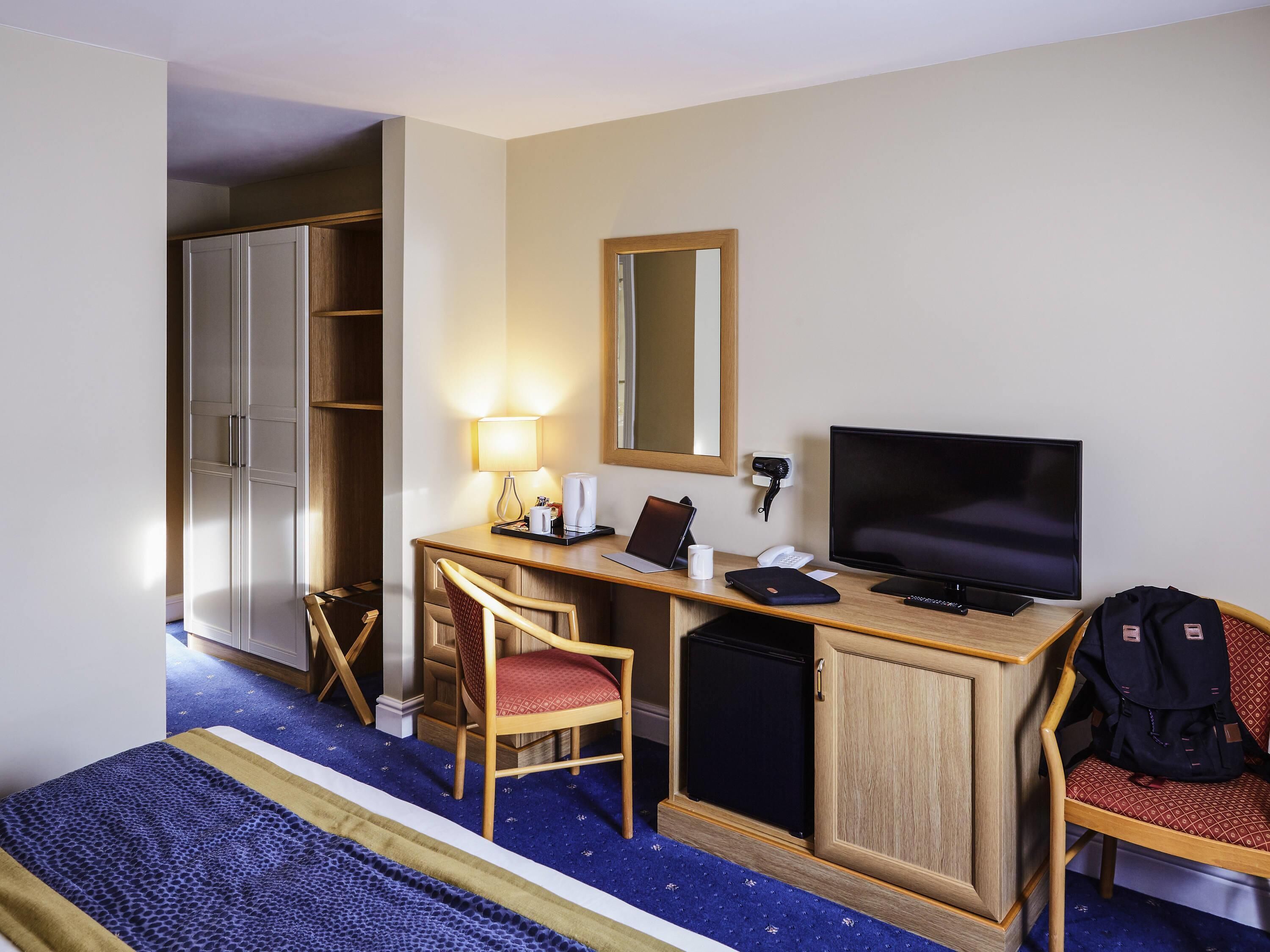 A hotel room with a bed, a desk with a computer, a chair, a wardrobe, and a television.