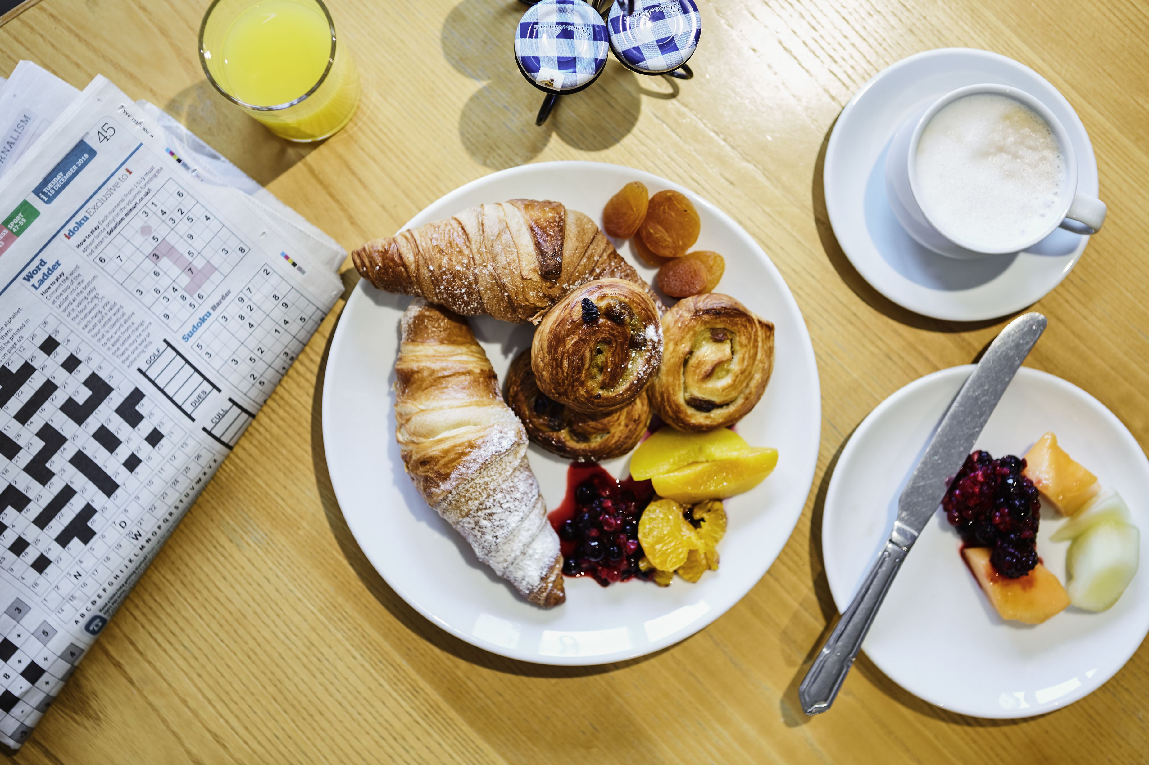 A breakfast setting with croissants, fruits, a cup of coffee, and a newspaper on a wooden table.