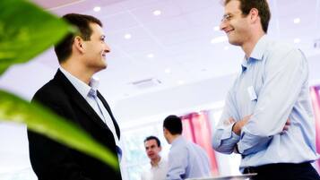 Two men in business attire conversing in a brightly lit indoor setting with a modern ceiling and visible windows.
