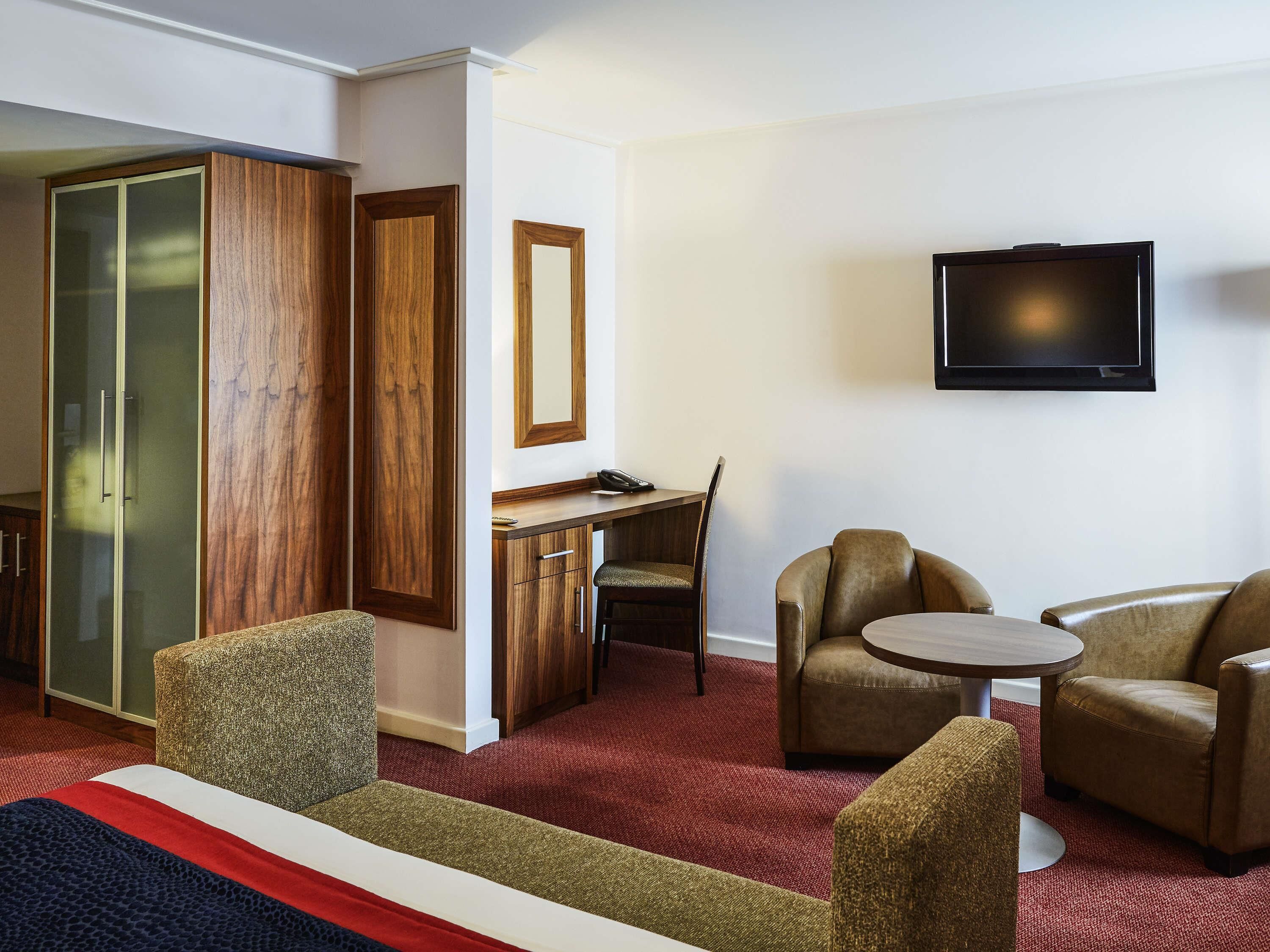A hotel room with a bed, a desk, two armchairs, a television, and a wardrobe.