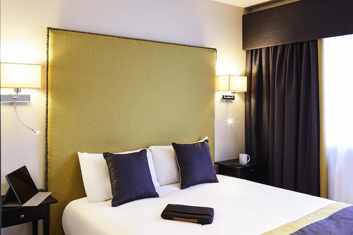 Una habitación de hotel con una cama grande, dos lámparas de noche, una mesita de noche con un ordenador portátil y una ventana con cortinas.
