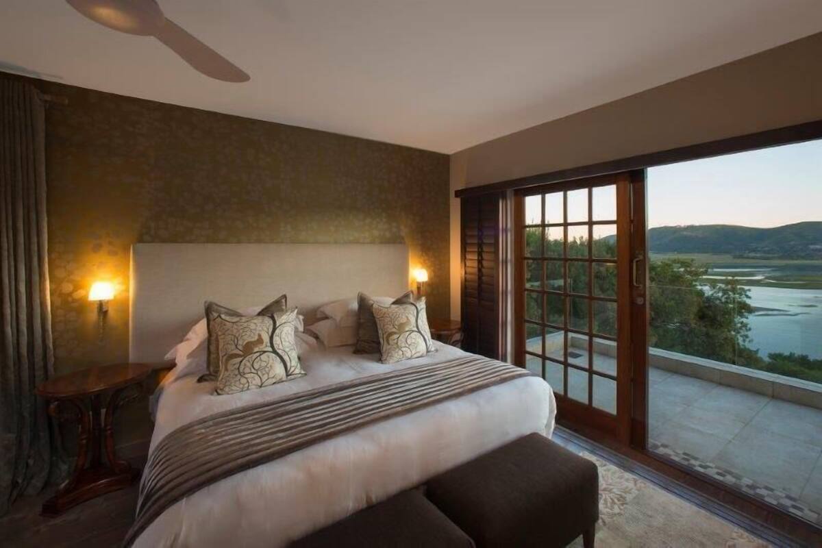 A bedroom with a large bed, two bedside lamps, a view of a lake through a sliding glass door, and a small table beside the bed.