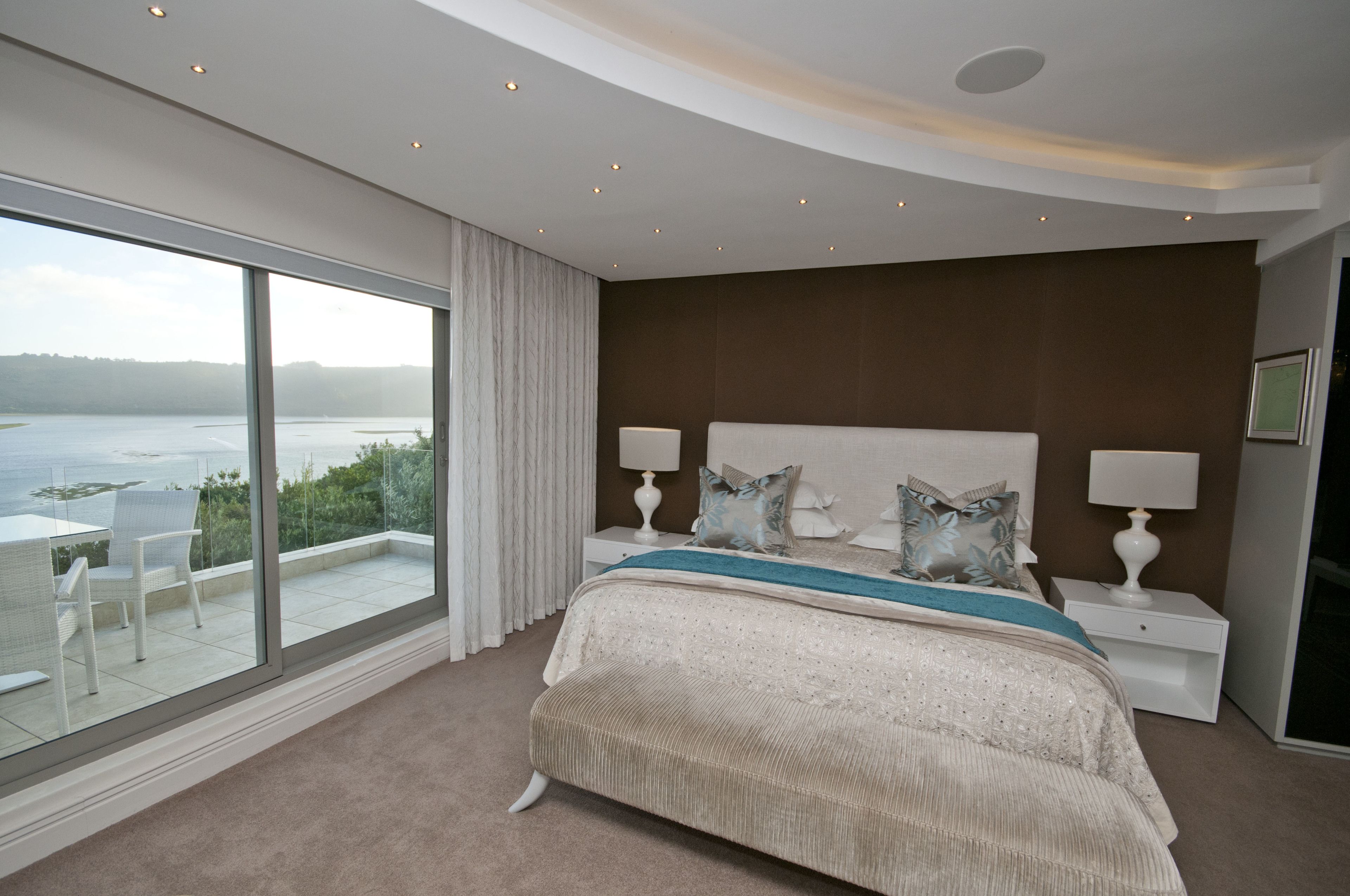 A bedroom with a large window, a bed with a blue accent, and a view of a body of water.
