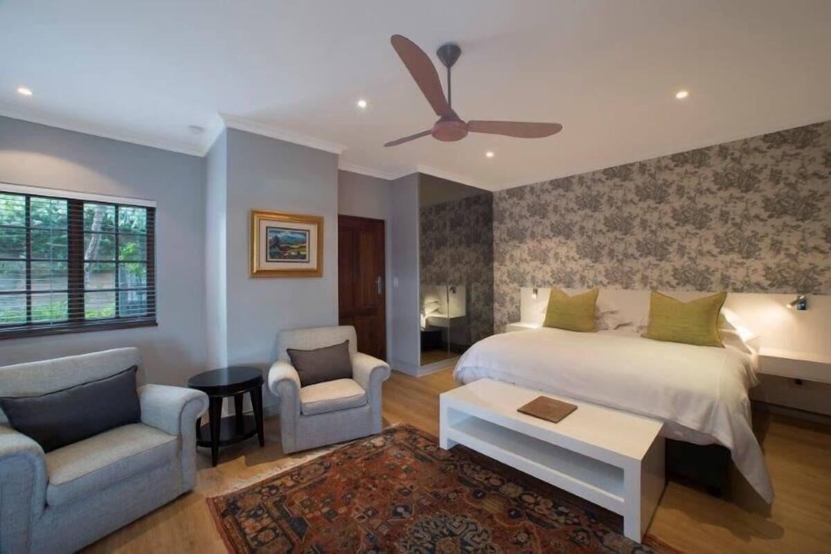 A bedroom with a bed, two armchairs, a small table, a ceiling fan, and a window with blinds.