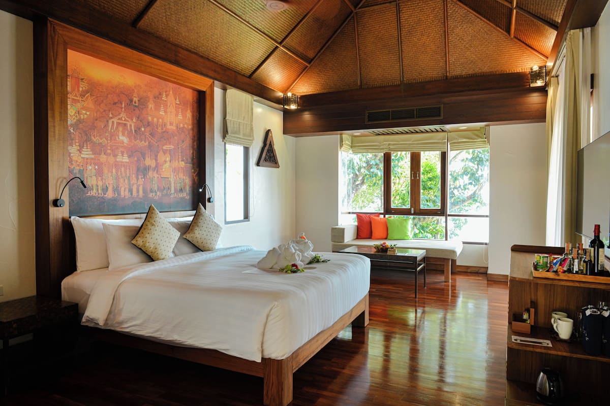 A large bed with pillows, a wooden headboard, a painting, a window with a view, and a small table with a chair.