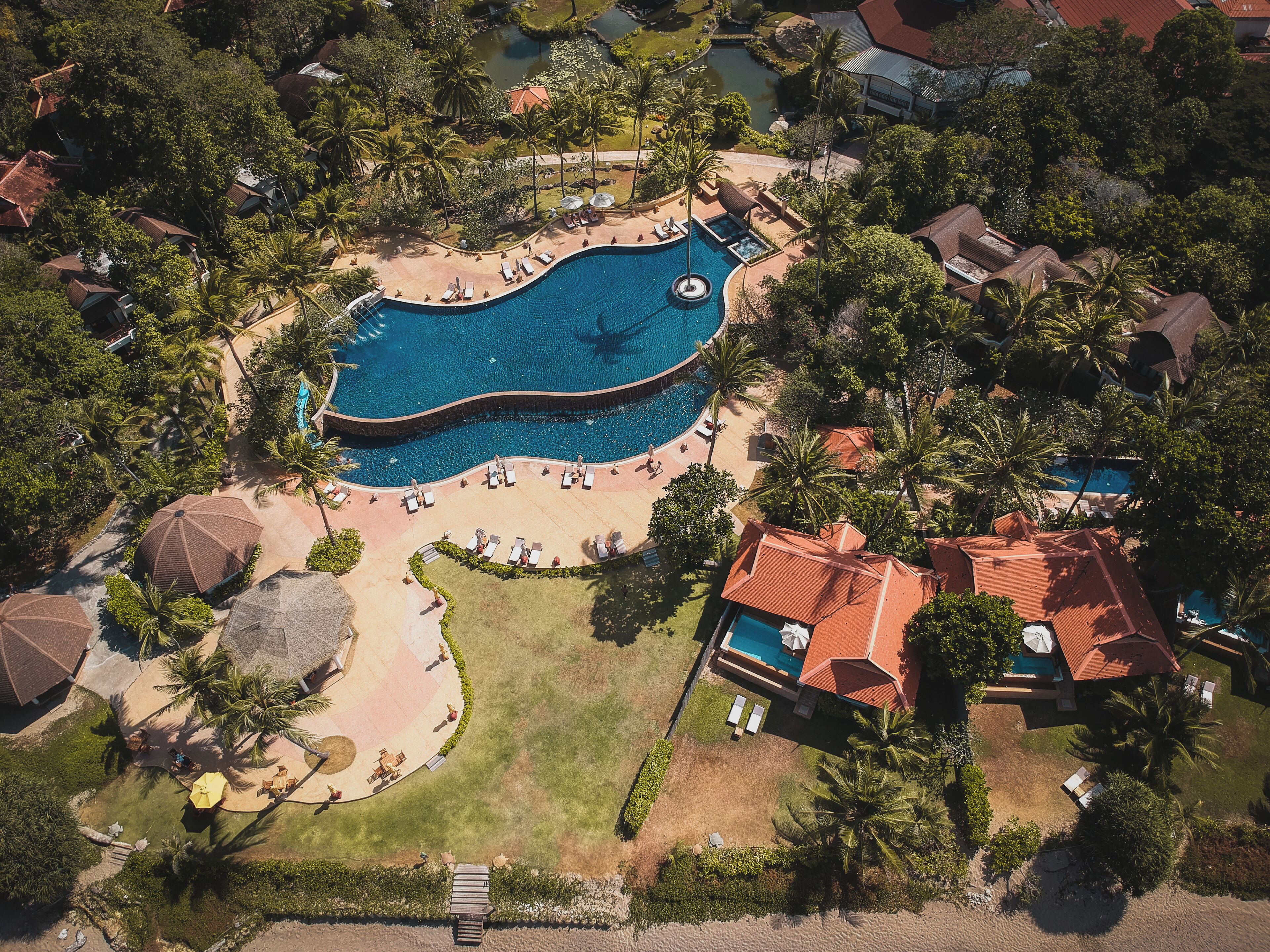 Vista aerea di un resort con una grande piscina, lettini prendisole e palme.