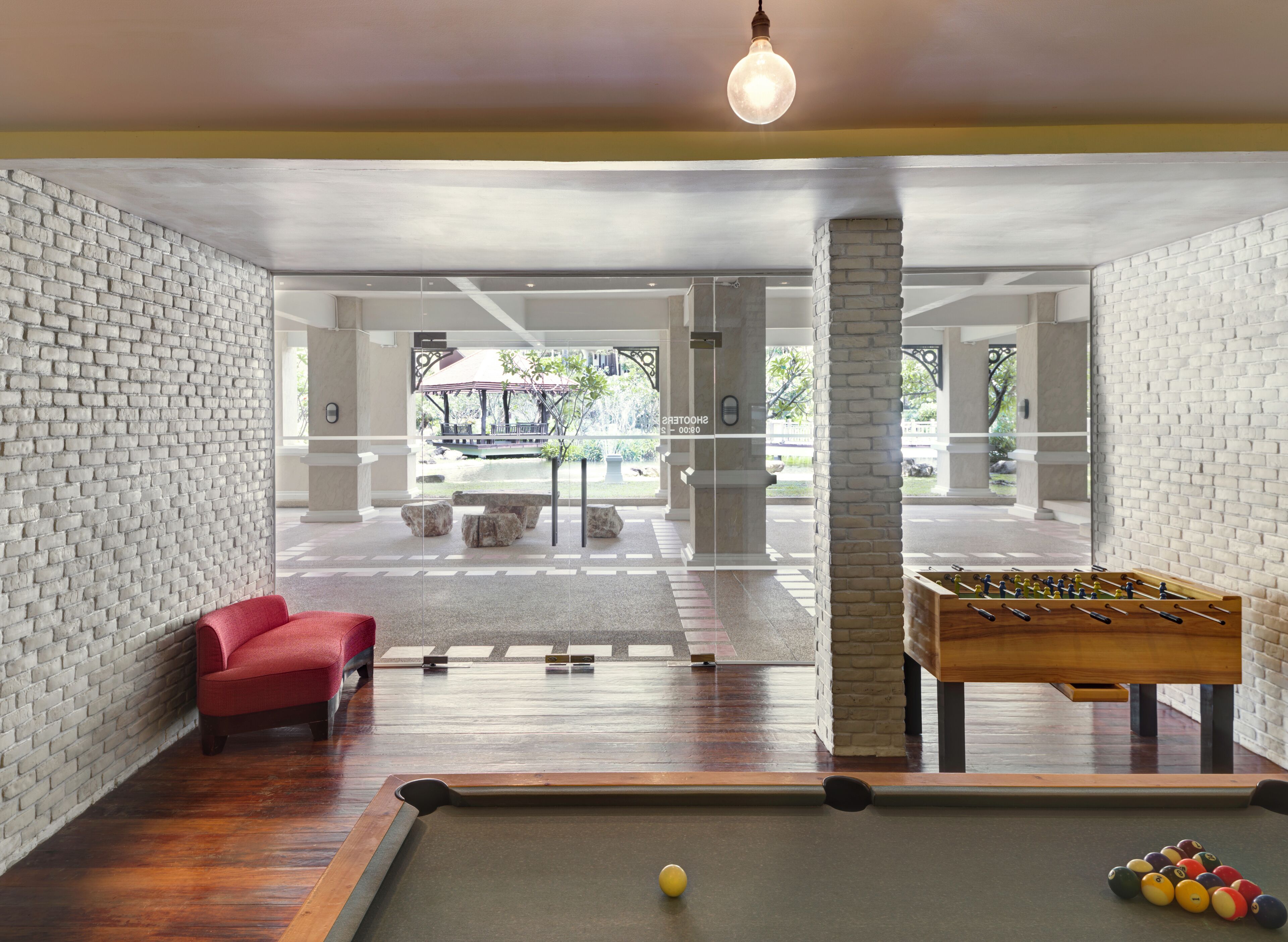 A pool table with a red chair and a foosball table in a room with brick walls and large windows.