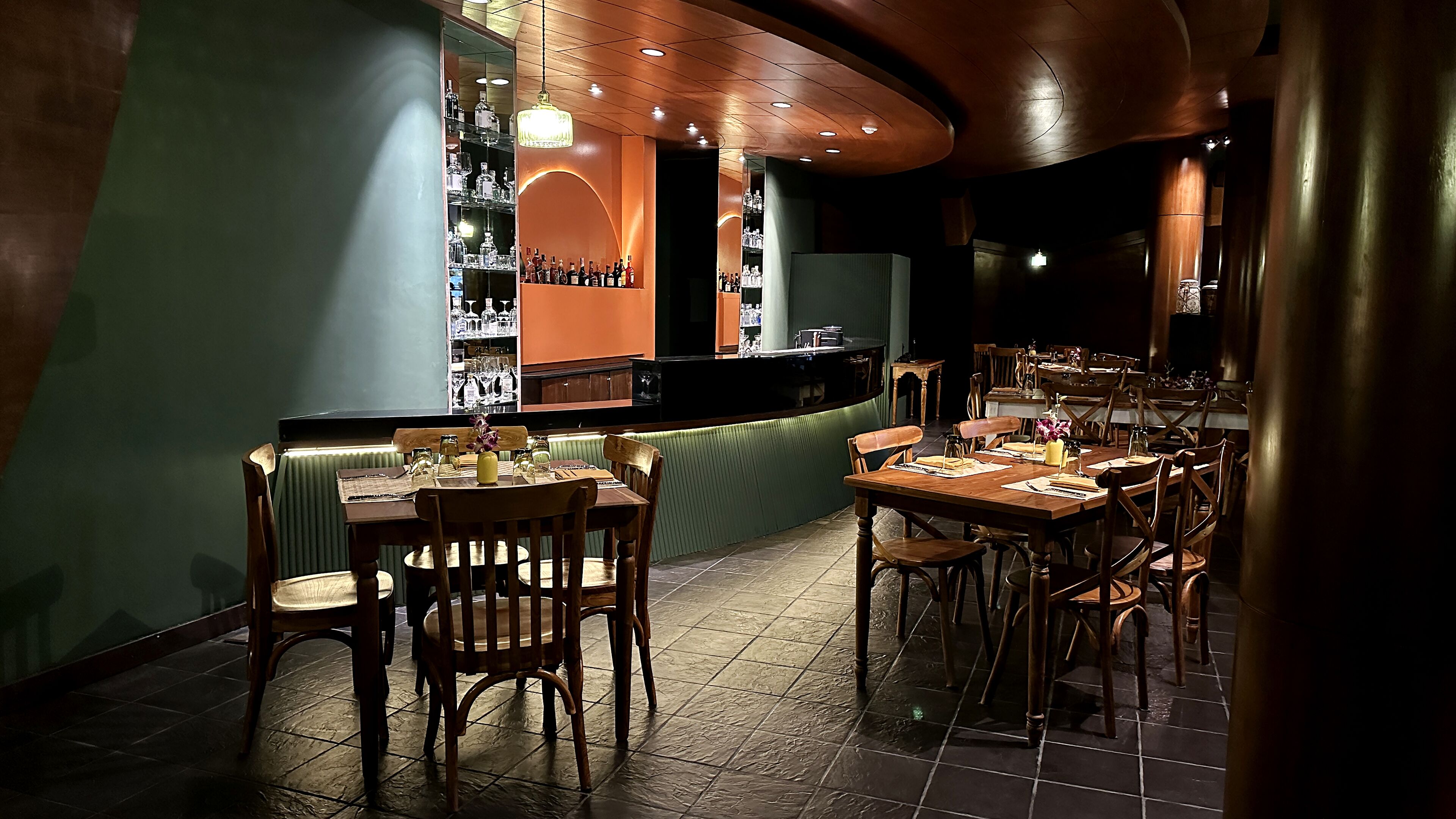 A restaurant with wooden tables and chairs, a bar area with shelves of bottles, and a green cushioned bench.