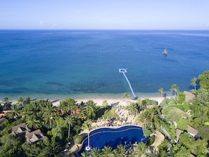 An aerial view of a resort with a large swimming pool, beachfront, and palm trees.