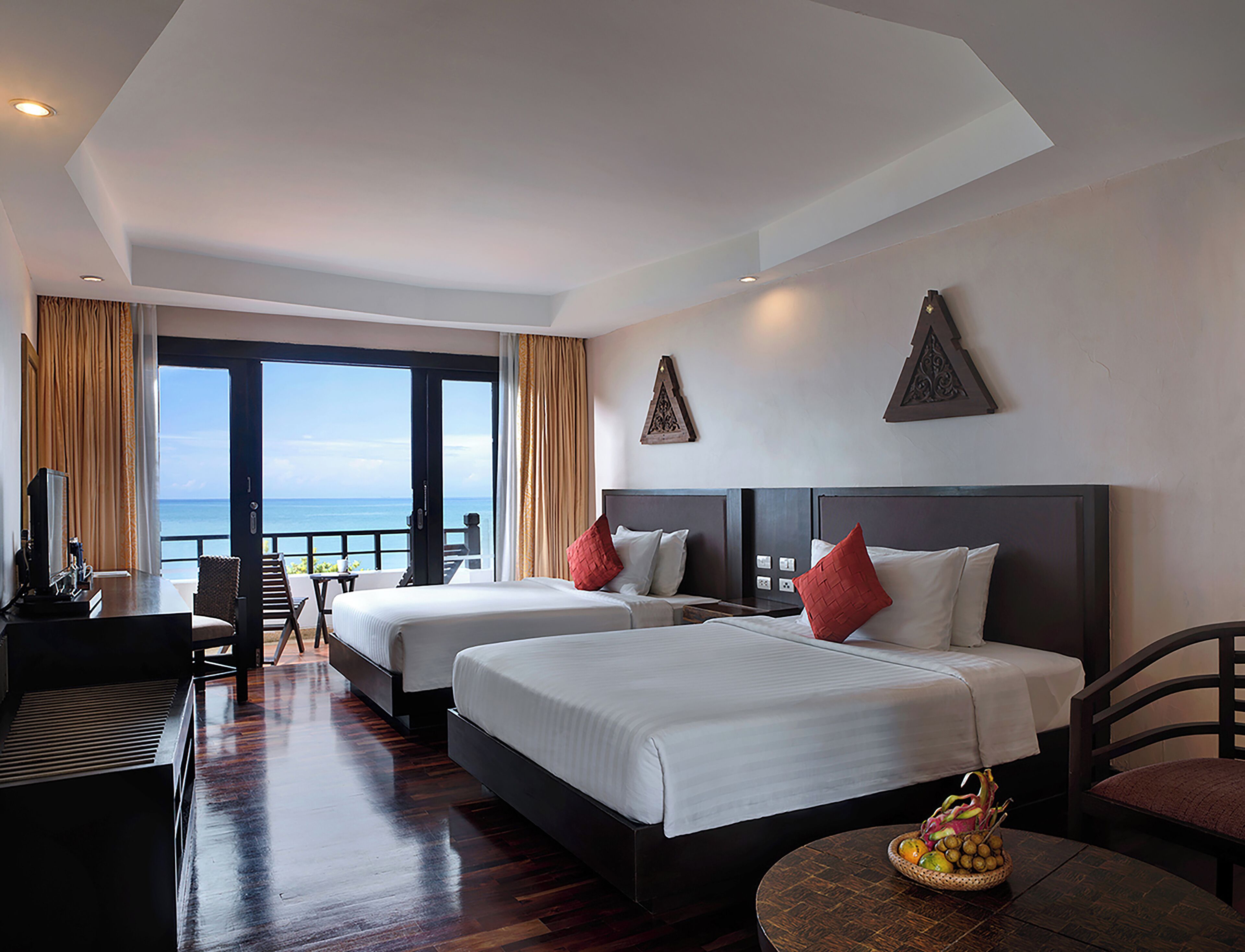 A hotel room with two beds, a desk, a chair, a small table with fruit, and a balcony with a view of the sea.