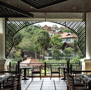 A covered outdoor seating area with tables and chairs, overlooking a hillside with traditional buildings and lush greenery.