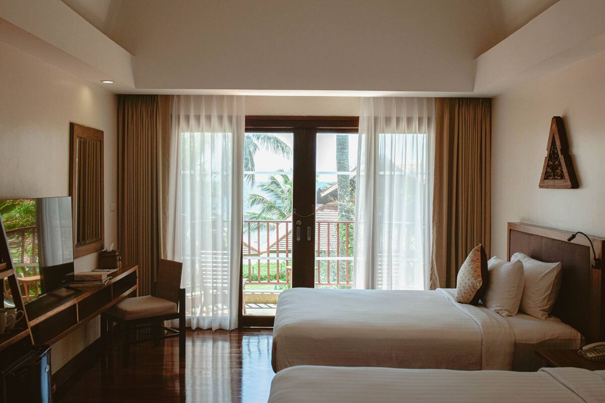 A hotel room with a bed, a desk with a computer, a chair, and a view of the outdoors through a large window.