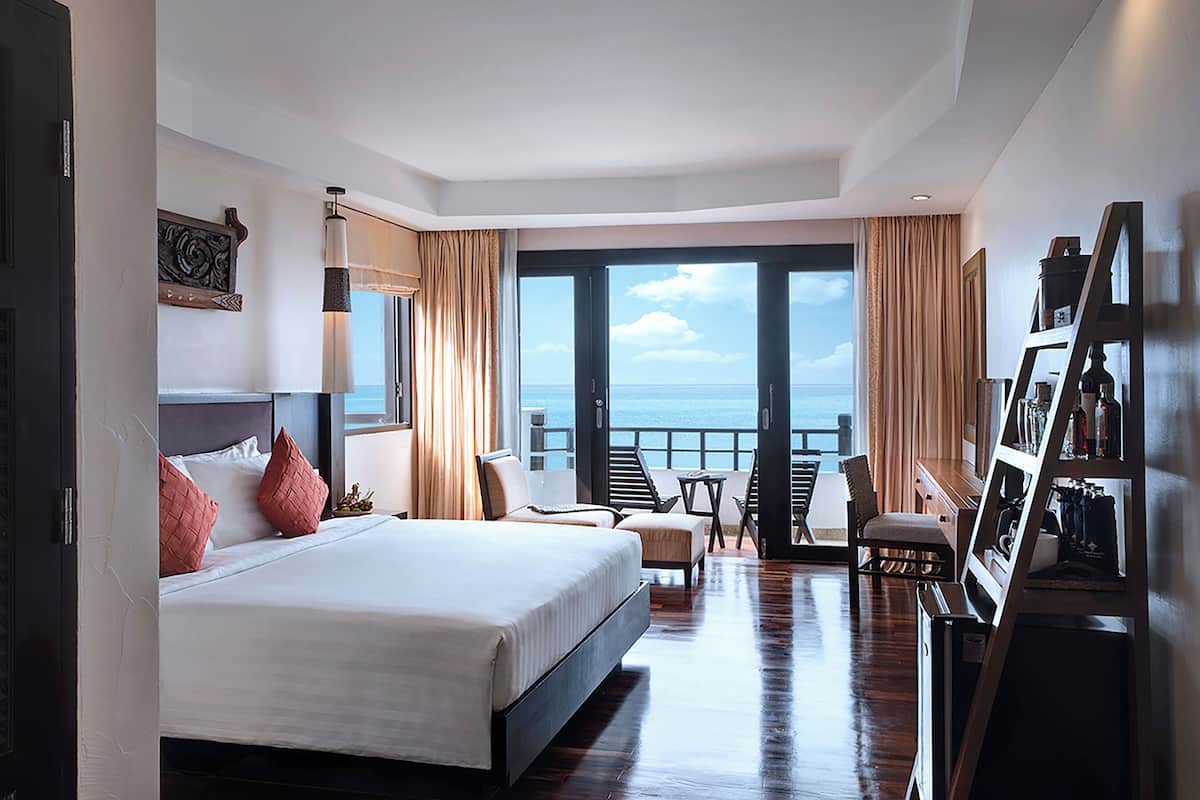 A hotel room with a large bed, a balcony with a view of the sea, a seating area with a table, and a wardrobe.