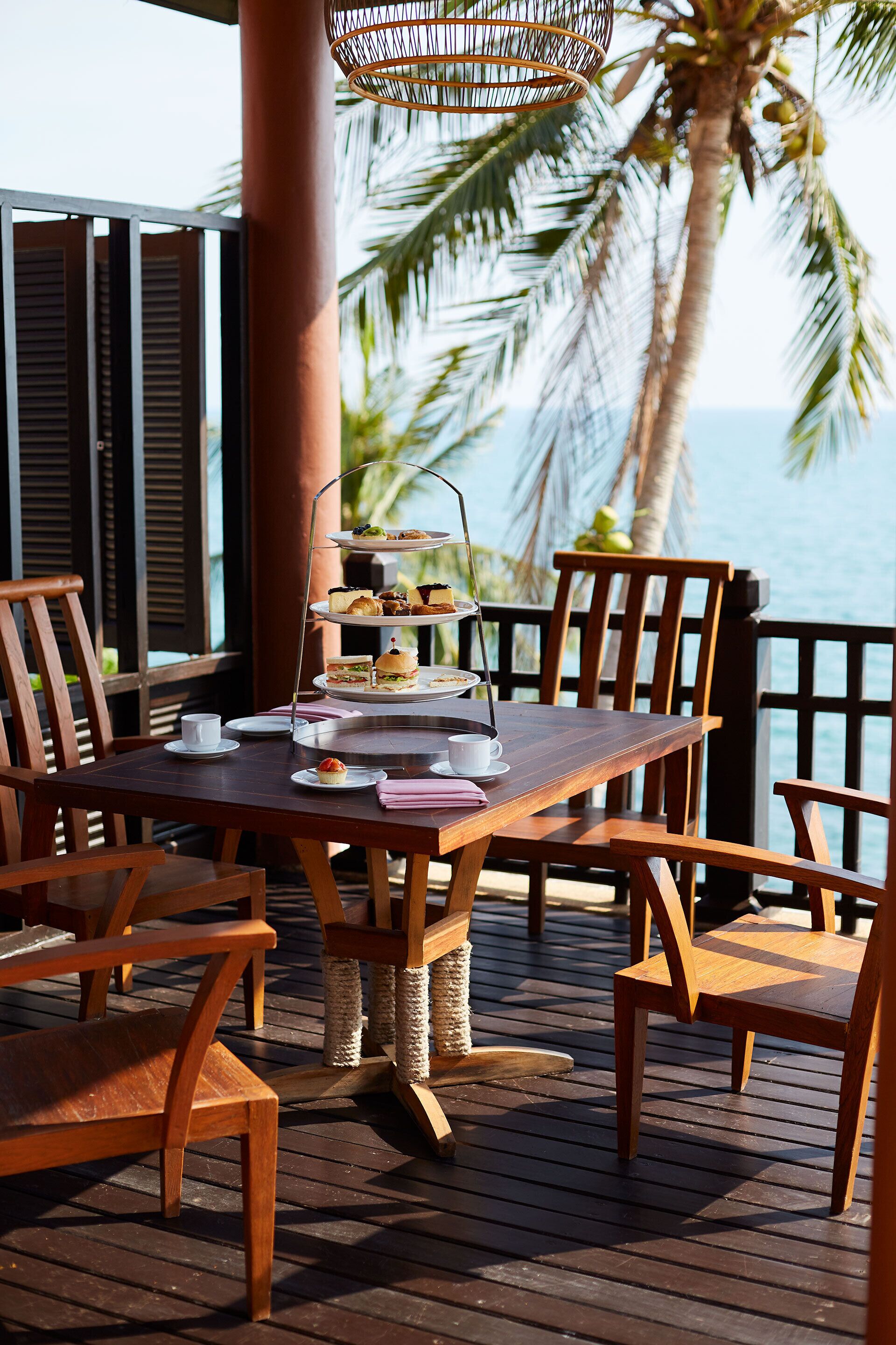 A wooden table with a tiered dessert stand, surrounded by wooden chairs on a wooden deck with a view of the ocean and palm trees.