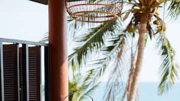 A wooden table with a tiered dessert stand, surrounded by wooden chairs on a wooden deck with a view of the ocean and palm trees.