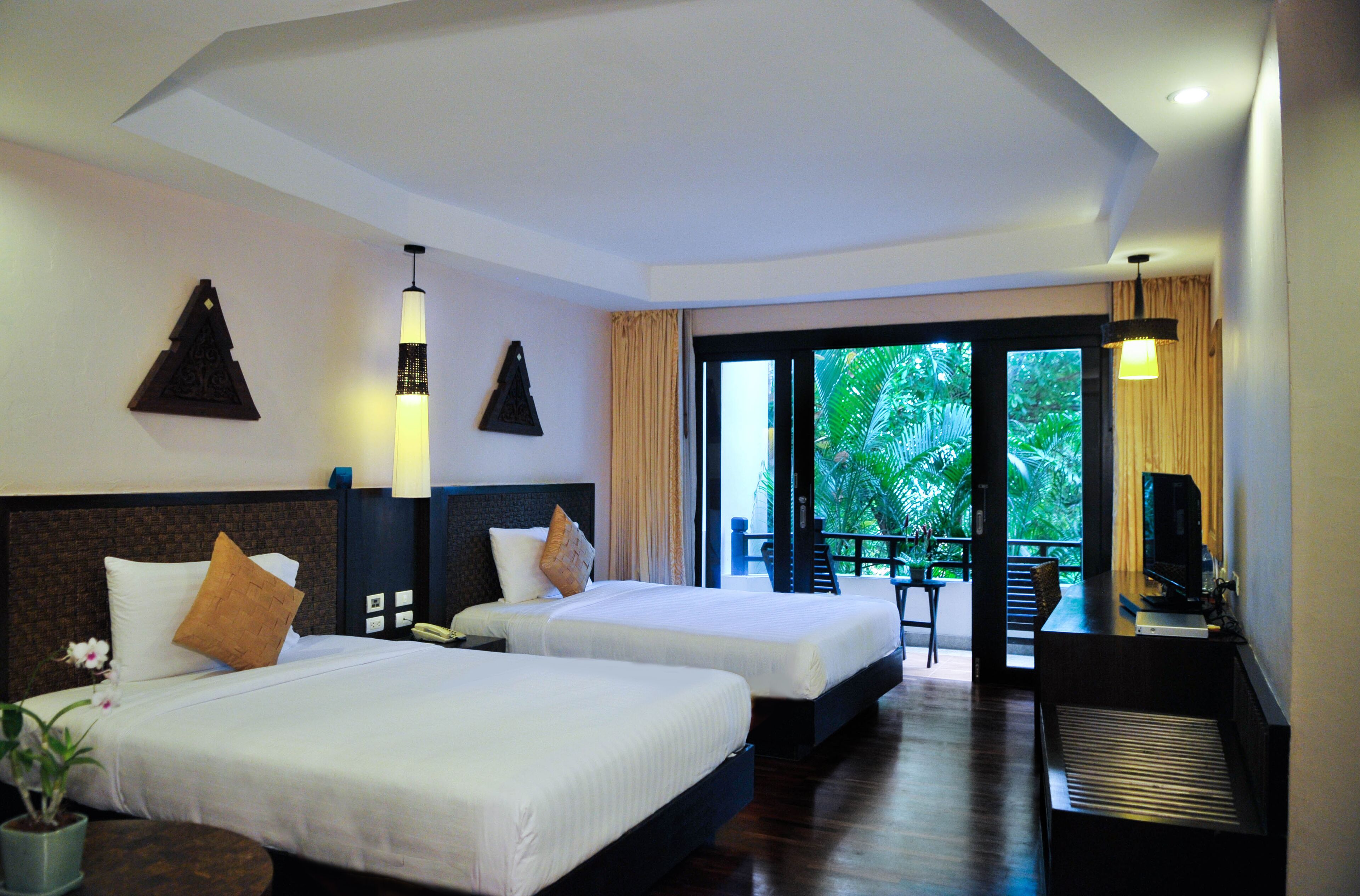 A hotel room with two beds, a desk, and a TV. There is a balcony with a view of greenery.