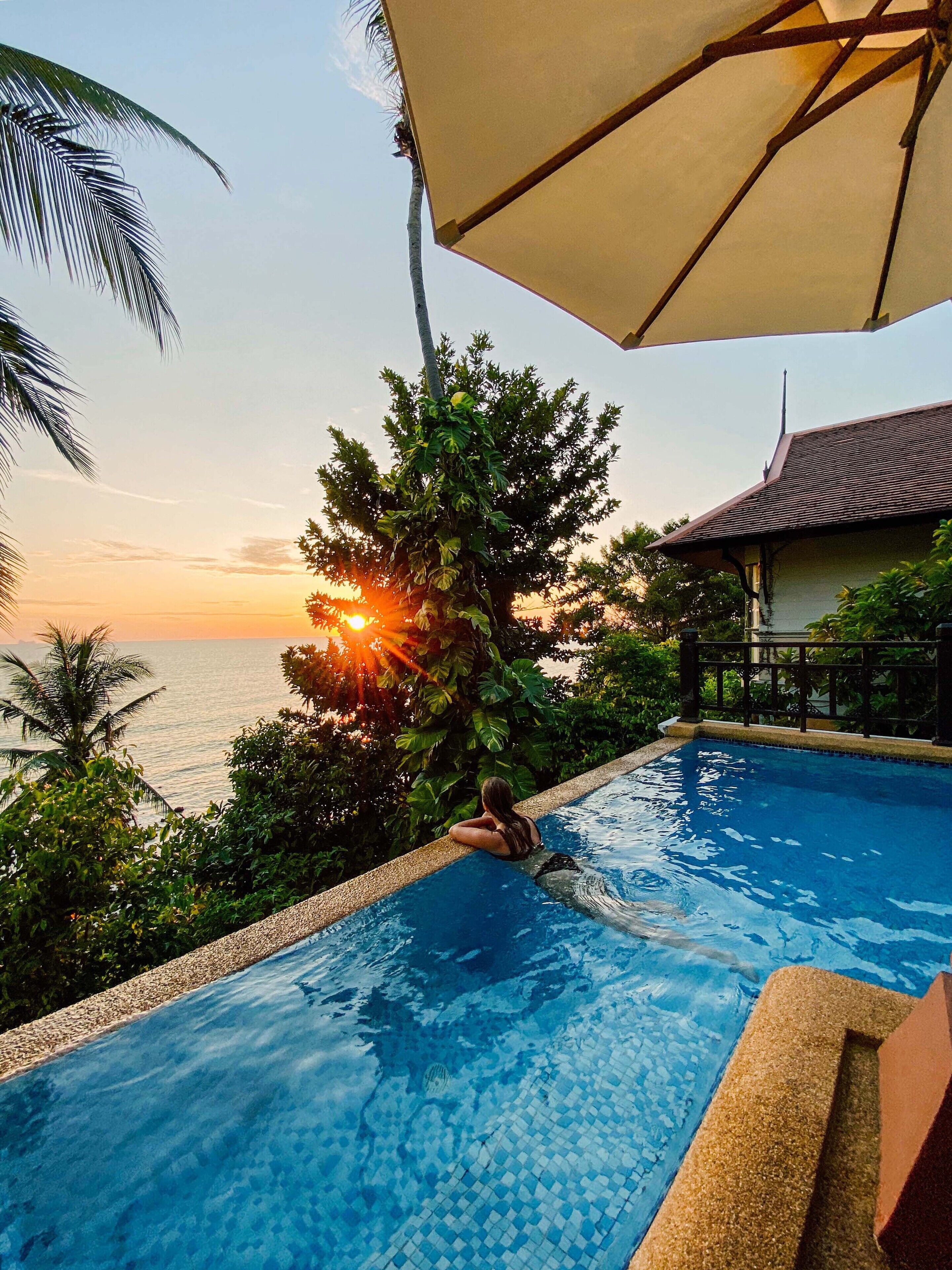 A swimming pool with a view of the sunset over the sea, surrounded by lush greenery and a traditional building.
