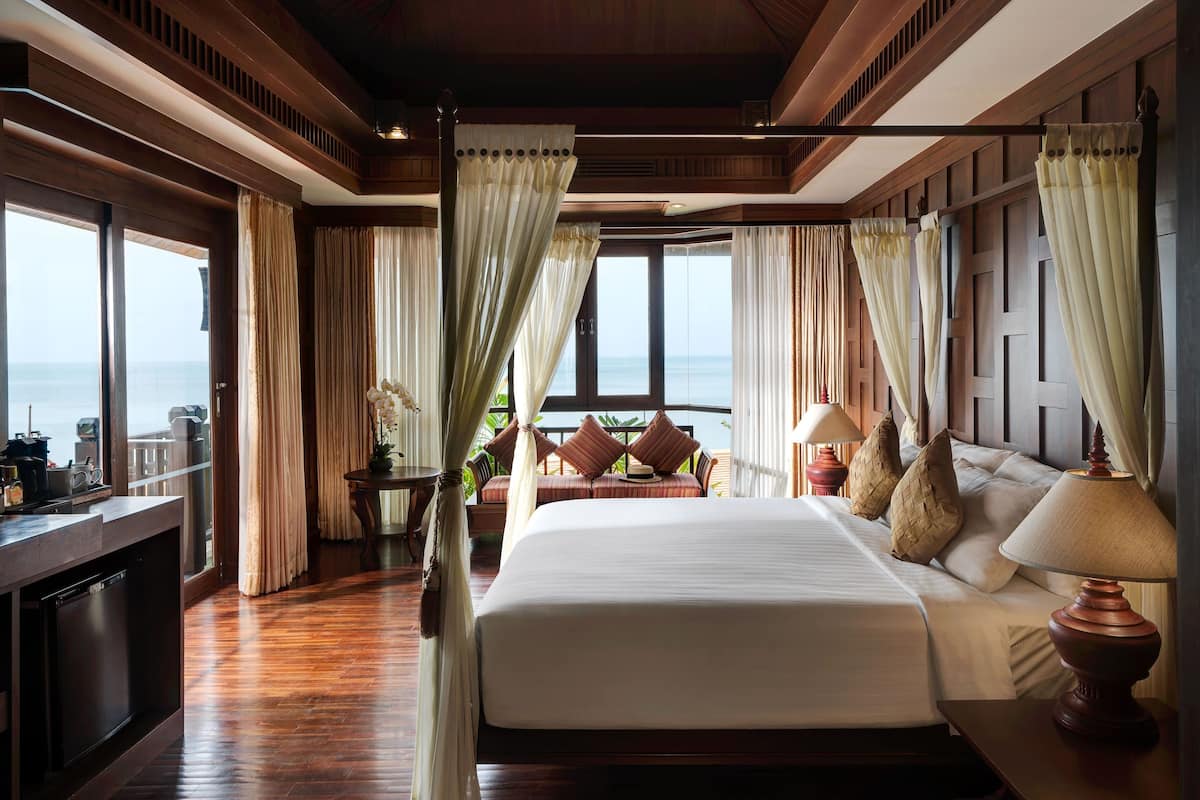 A spacious bedroom with a large bed, a bedside table with a lamp, and a view of the ocean through large windows.