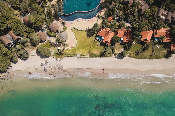 An aerial view of a coastal property with a beach, a swimming pool, and several houses with red roofs.