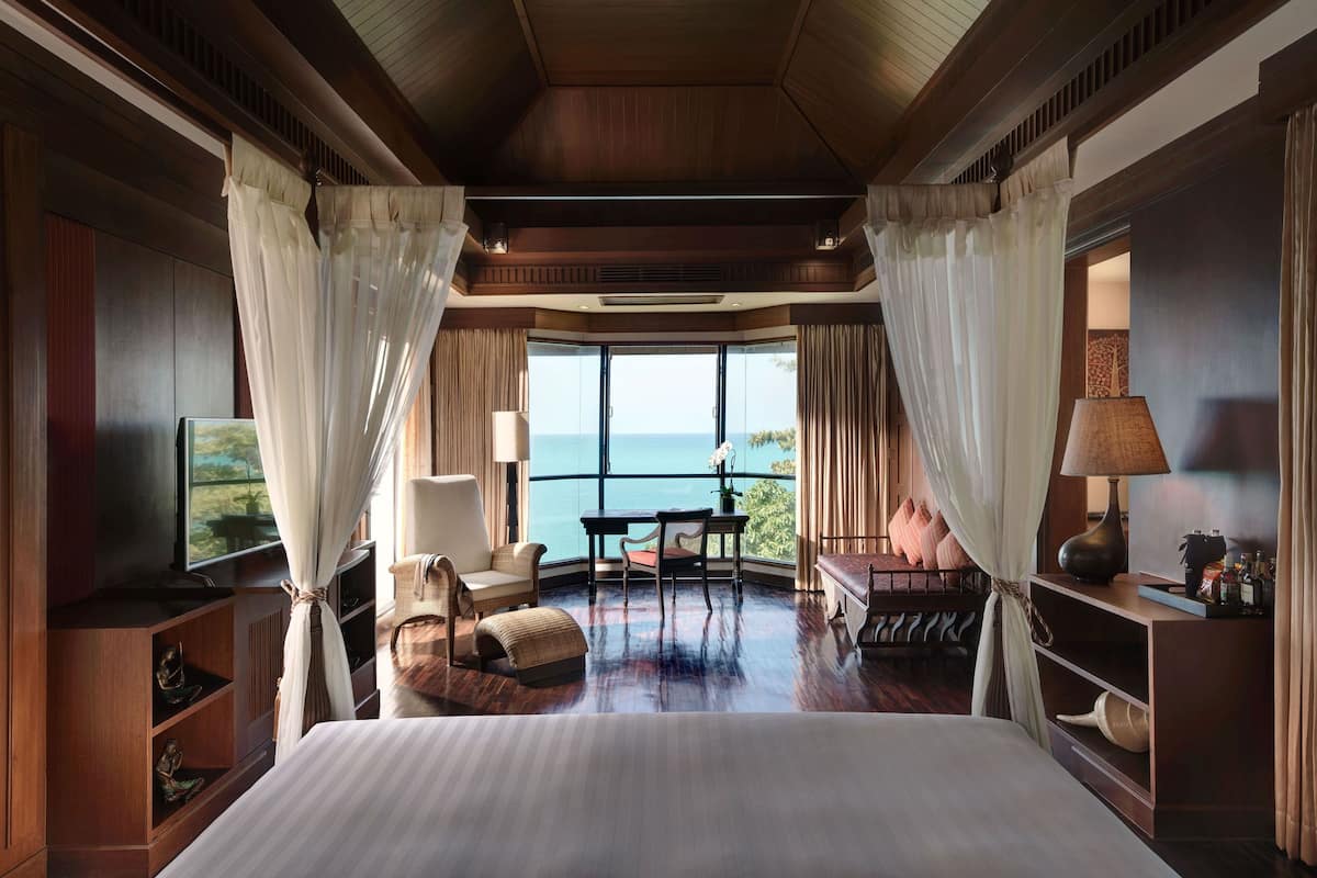 A spacious bedroom with a large bed, a television, a seating area with a table and chairs, and a view of the ocean through large windows.