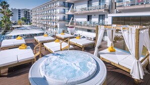 Terraza en la azotea con jacuzzi, rodeada de sillones y camas sol, con vistas a una piscina y un edificio con balcones.