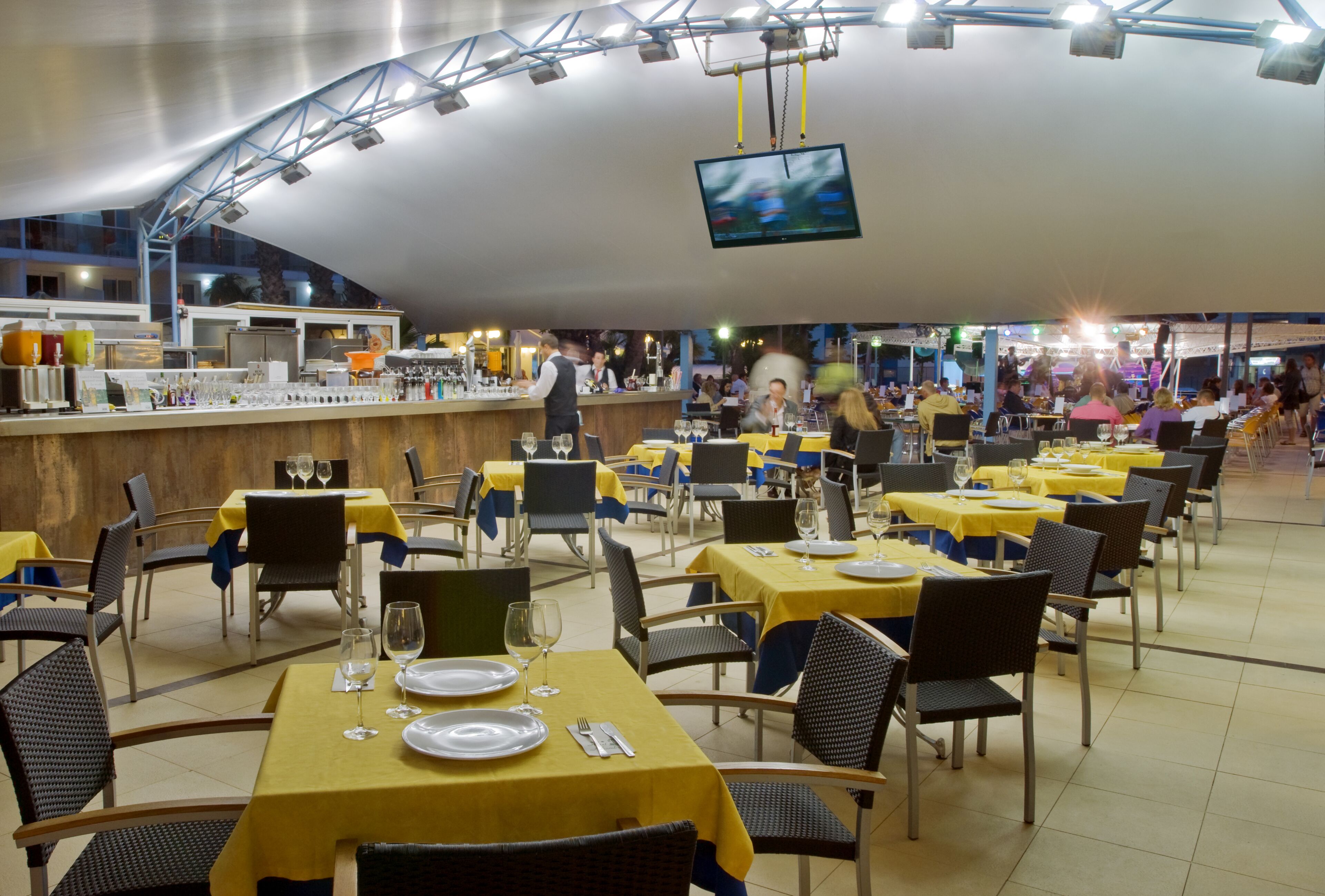 Un restaurante espacioso con varias mesas y sillas, una zona de bar y una pantalla grande que muestra una imagen desenfocada.