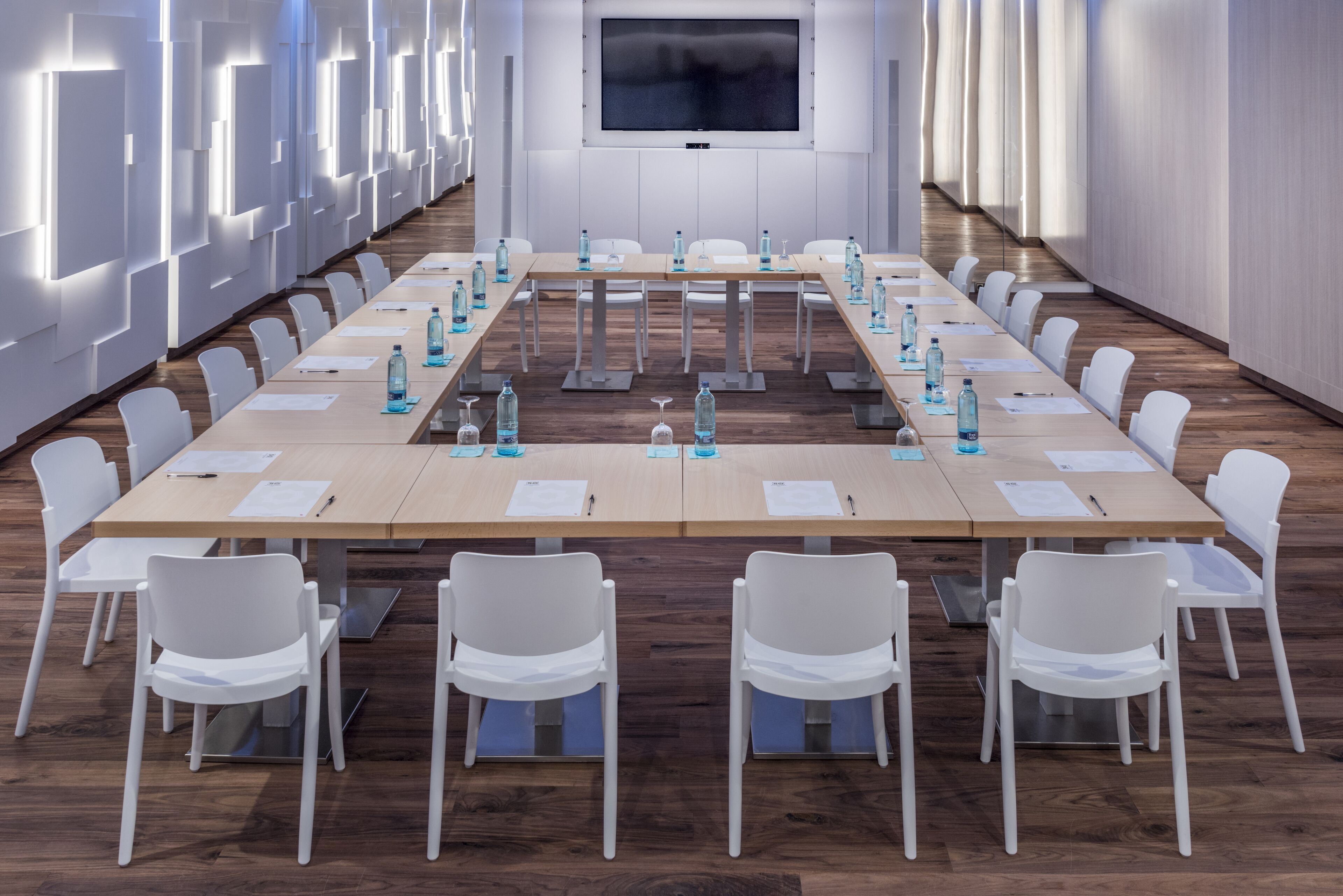 Un salón de conferencias con una larga mesa, sillas blancas, botellas de agua y documentos.